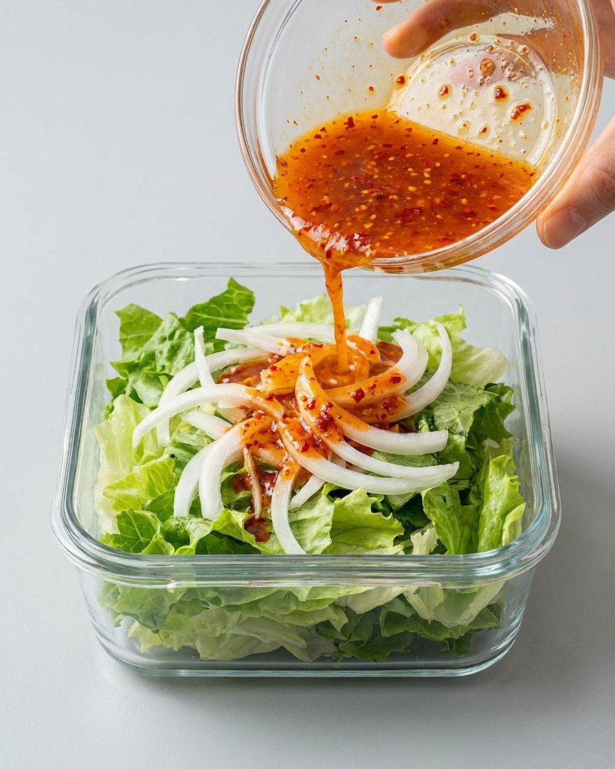 Brodo di kimchi rosso e piccante versato su lattuga e cipolle in un contenitore rettangolare di vetro.