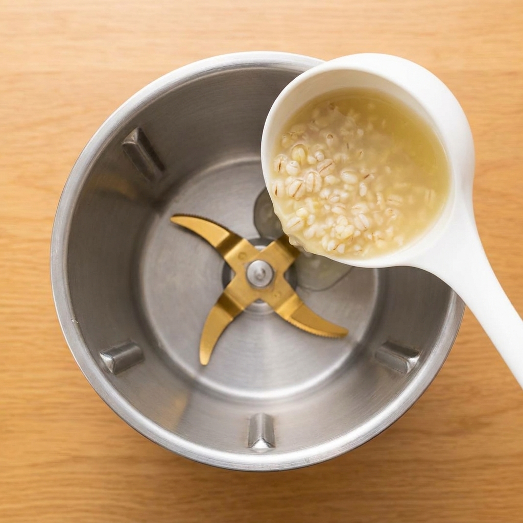 Un mestolo bianco che versa un porridge dorzo denso e chiaro nel bicchiere di un frullatore in acciaio.