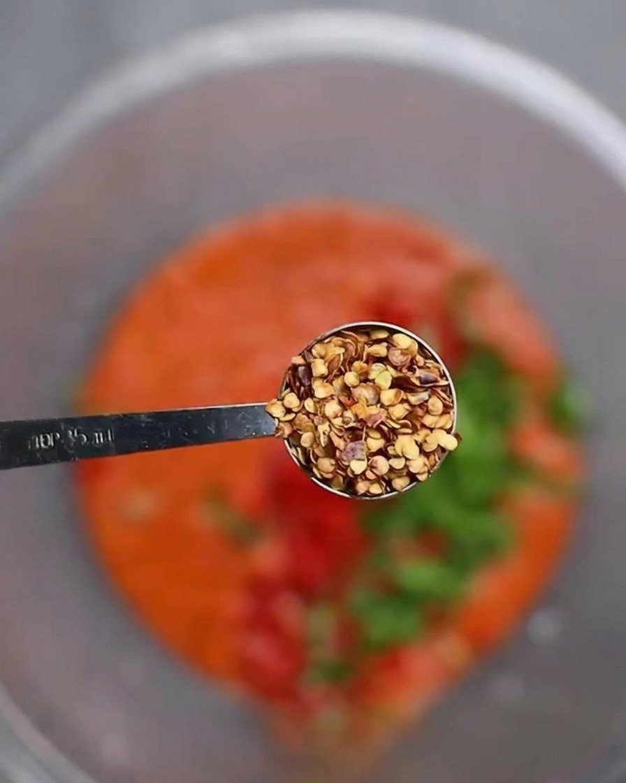 Cucchiaio dosatore con semi di peperoncino sopra una ciotola con pasta rossa e verdure tritate.
