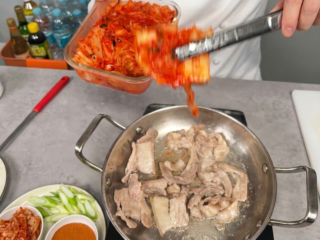 Una porzione abbondante di kimchi rosso versata da un contenitore in una padella con pancetta rosolata.