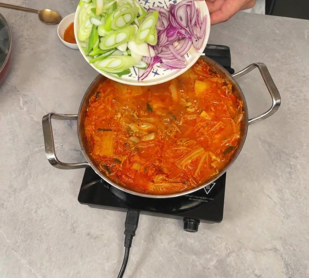 Cipollotti e cipolla rossa affettati che scivolano da un piatto in uno stufato di kimchi rosso bollente.