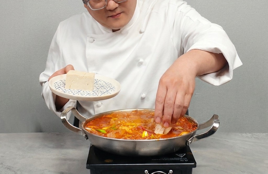 Una persona che adagia fette di tofu bianco compatto in una pentola di stufato di kimchi bollente.