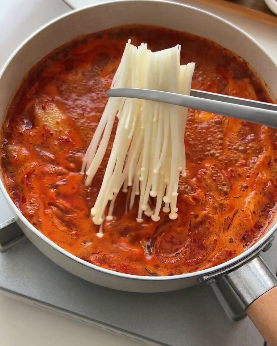 Pinze che adagiano un mazzetto di funghi enoki freschi nel brodo rosso bollente.
