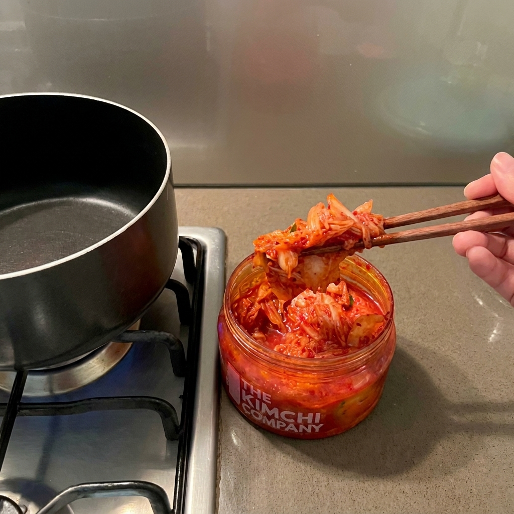 Una mano usa bacchette di legno per prelevare il kimchi rosso da un barattolo sopra una pentola nera vuota.