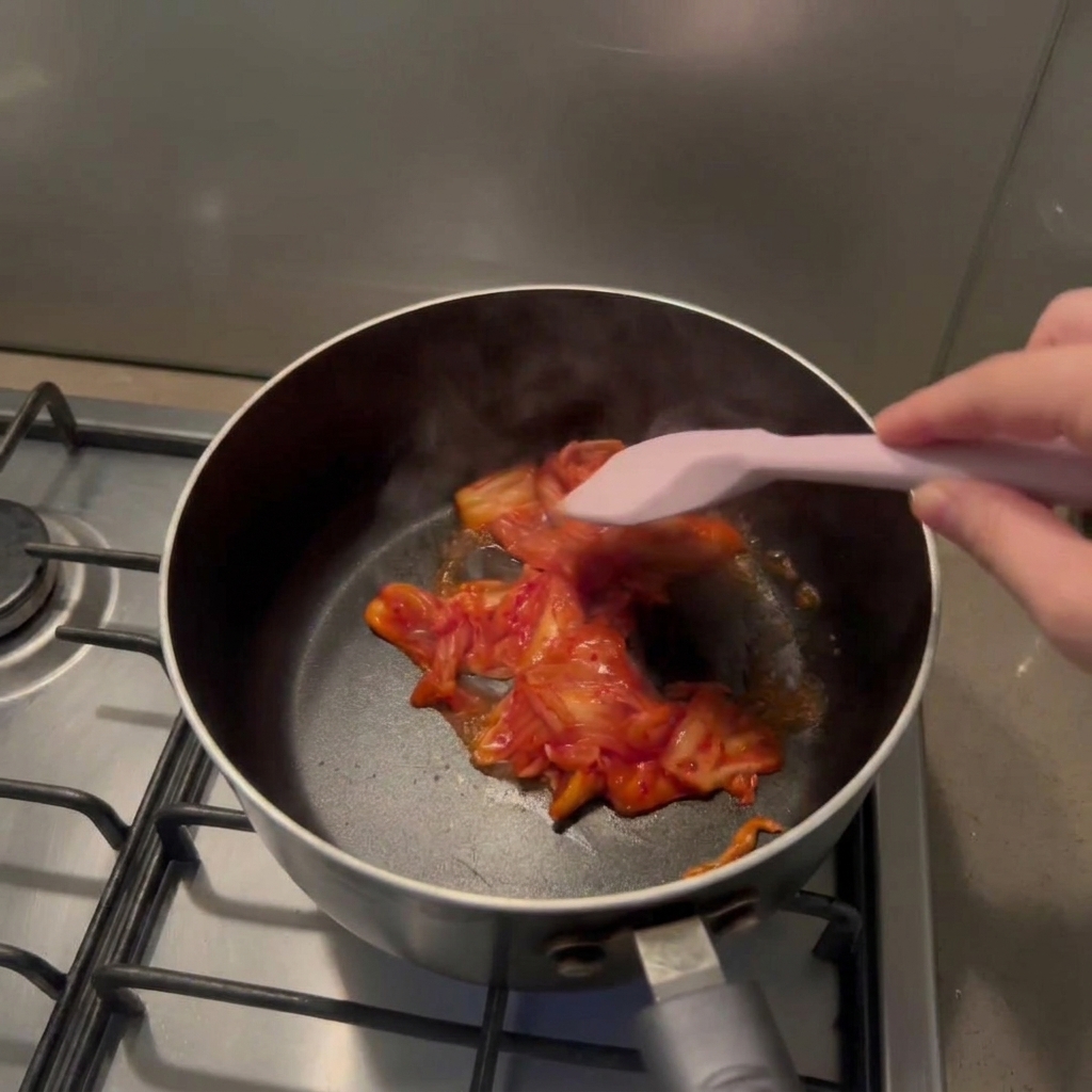 Una mano usa una spatola di silicone rosa per soffriggere il kimchi rosso tritato in una pentola nera.