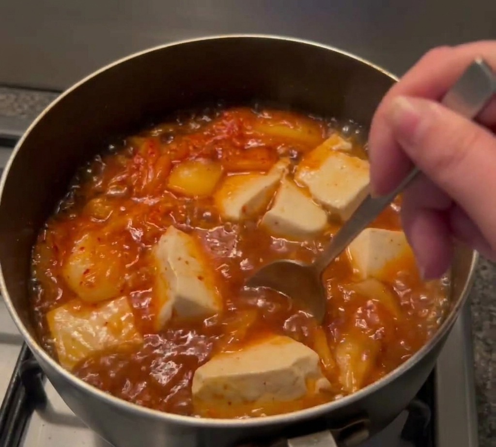 Un cucchiaio di metallo mescola la pasta di peperoncino in una pentola di zuppa con tofu e kimchi.
