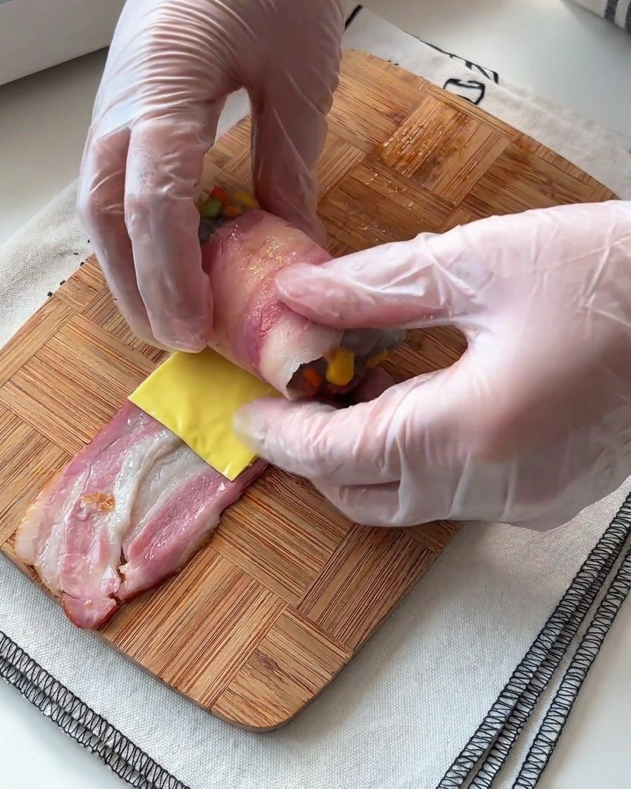 Mani che arrotolano strettamente il bacon attorno al ripieno di gamberi e formaggio su una superficie di legno.