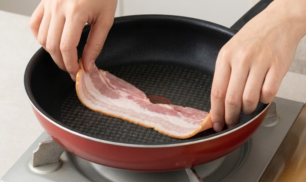 Mani che adagiano fette di bacon crudo in una padella antiaderente calda e leggermente unta.