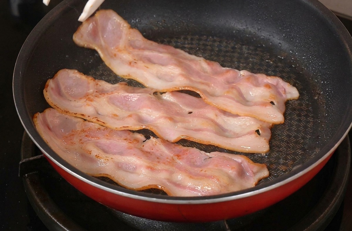Pinze da cucina che girano fette di bacon arricciate in una padella rossa sul fornello.