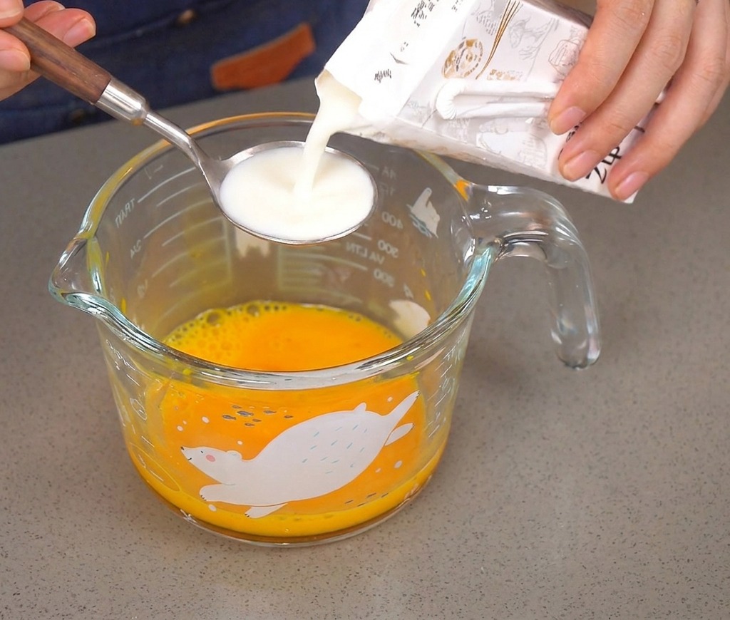 Latte versato da un cartone in un cucchiaio dargento sopra una ciotola con uova sbattute.