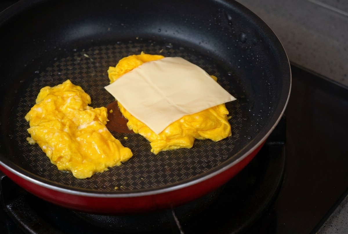 Padella antiaderente con due mucchietti di uova strapazzate su cui viene adagiata una fetta quadrata di formaggio chiaro.