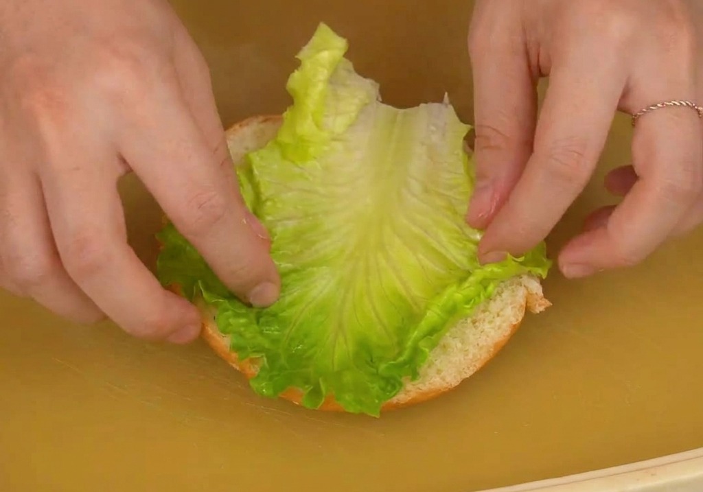Due mani che posizionano con cura una foglia di lattuga fresca sulla metà inferiore di un panino da burger su un tagliere.