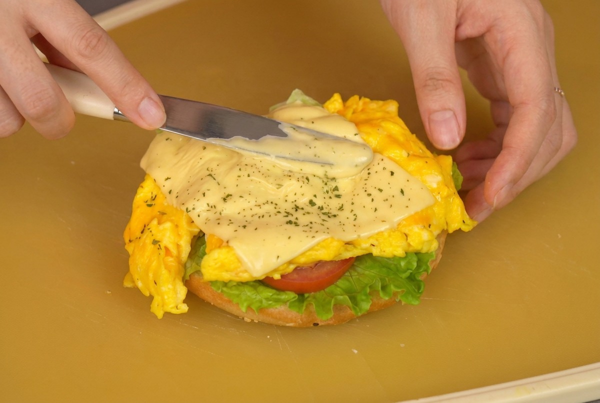 Primo piano di una persona che usa una spatola per trasferire uova strapazzate e formaggio su un panino con lattuga e pomodoro.
