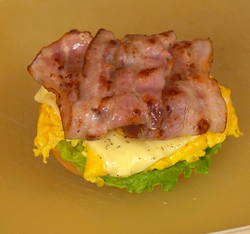 Burger aperto su un tagliere che mostra strati di lattuga, pomodoro, uova al formaggio e bacon croccante.