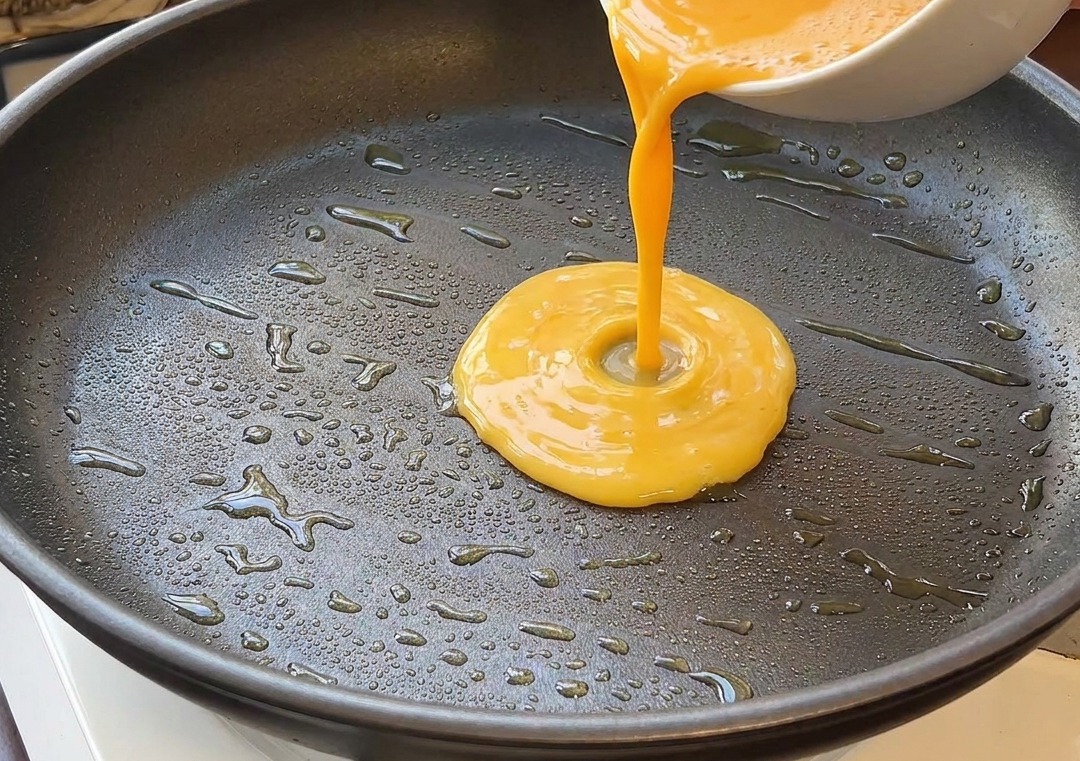 Composto di uova sbattute versato da una ciotola bianca in una padella nera con olio caldo.
