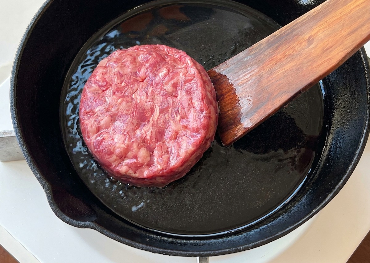 Un hamburger di carne cruda adagiato in una padella di ghisa calda con una spatola di legno.