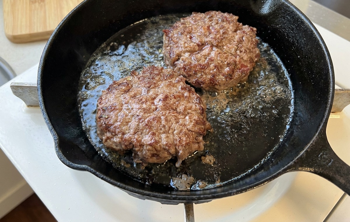 Due hamburger che sfrigolano in padella, mostrando una crosticina ben brunita e caramellata.