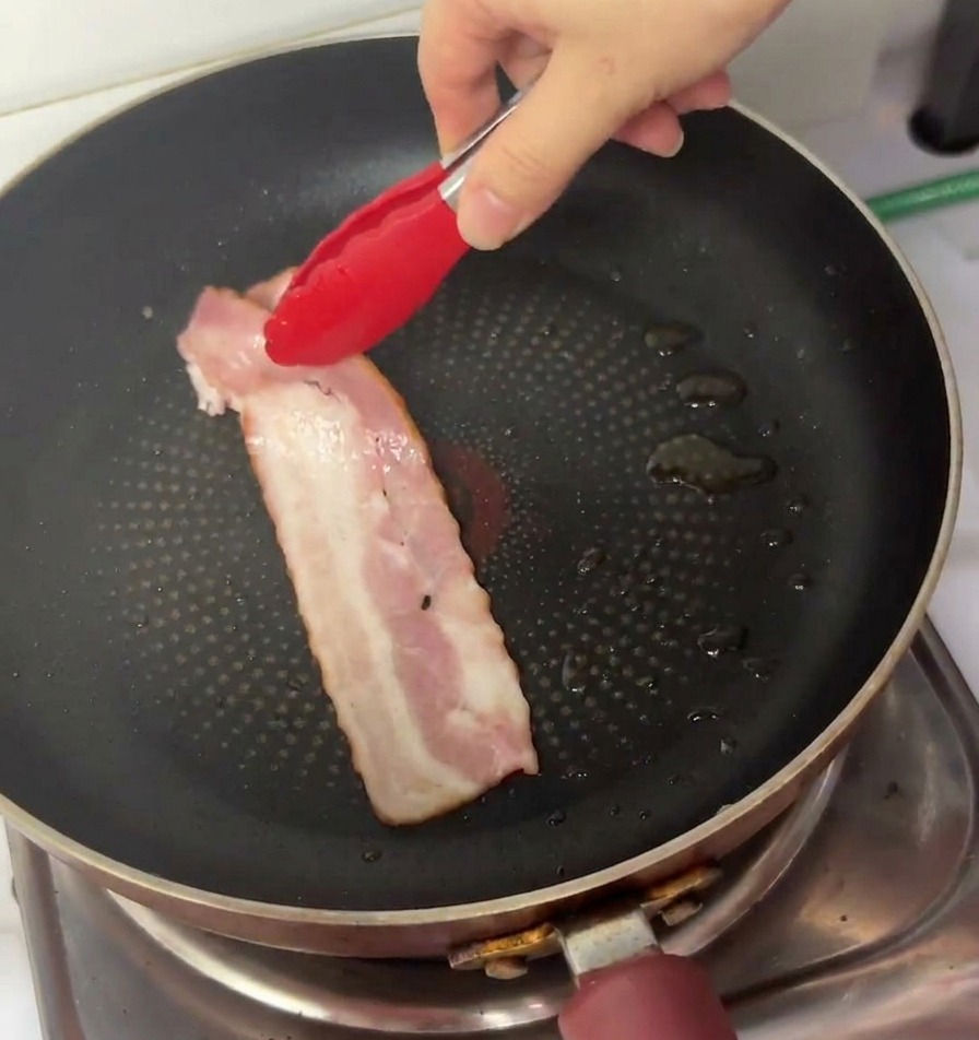 Una mano usa pinze con punte rosse per mettere una striscia di bacon crudo in una padella antiaderente.