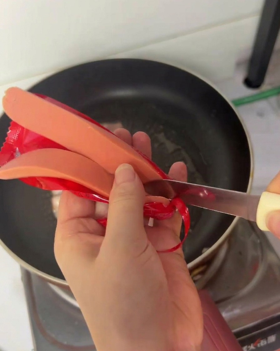 Una persona usa un coltellino per tagliare a metà longitudinalmente un wurstel sopra una padella.