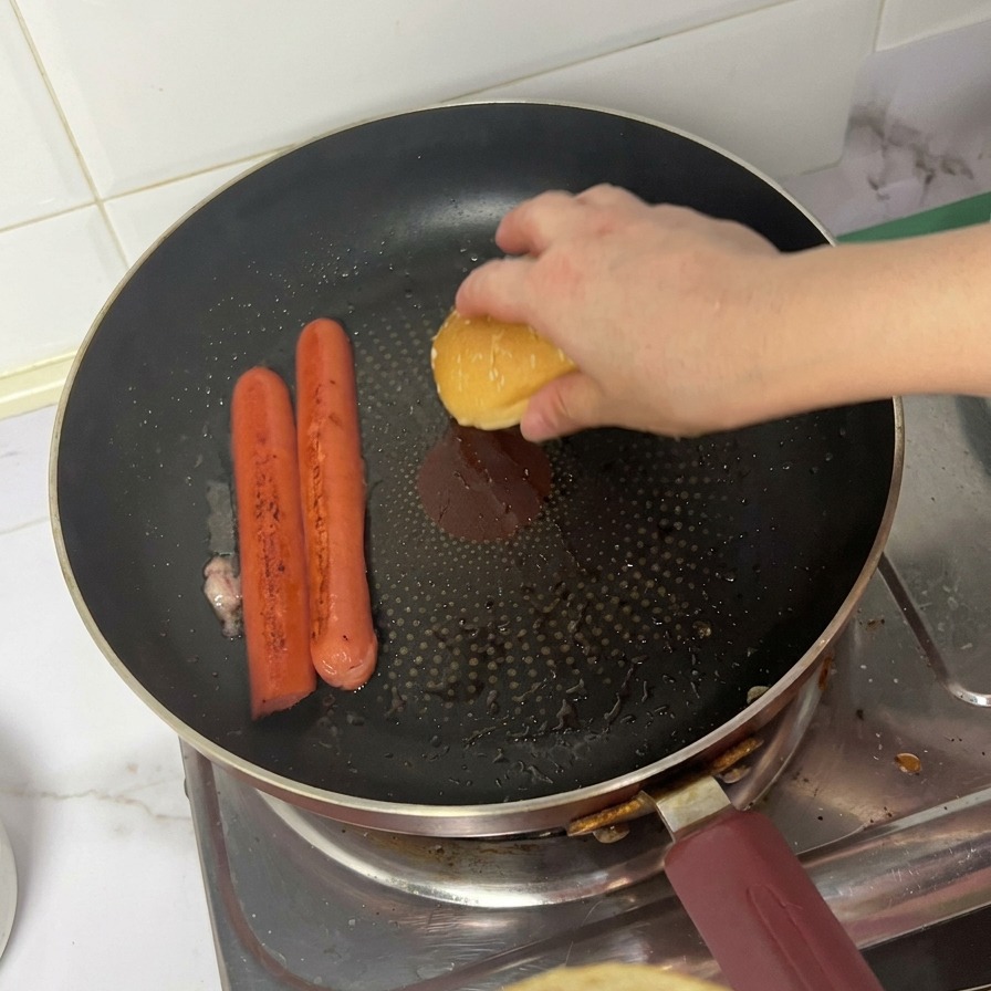 Una persona appoggia la parte superiore di un panino al sesamo a faccia in giù in padella accanto ai wurstel in cottura.