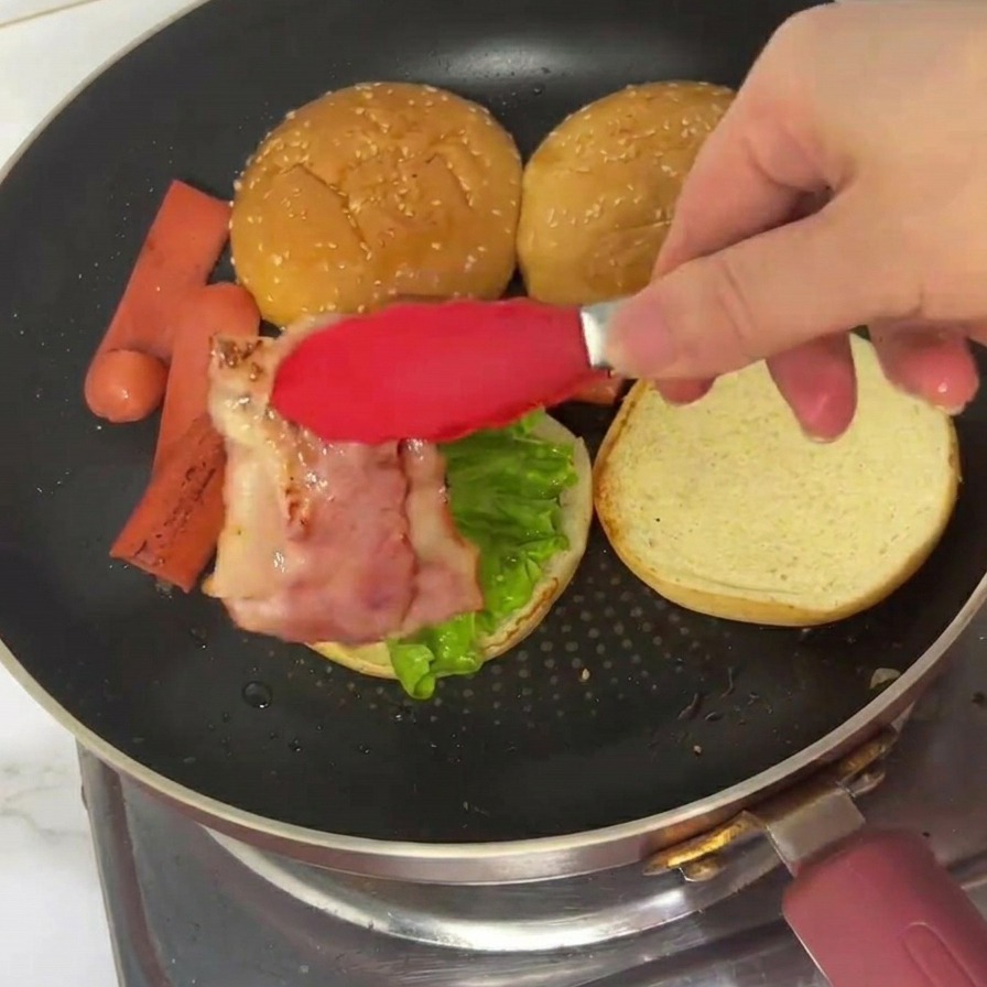Pinze rosse che posizionano una striscia di bacon cotto e ondulato su un panino già farcito con lattuga.
