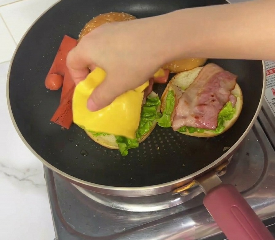 Una mano che mette una fetta quadrata di formaggio giallo su un burger in fase di assemblaggio in padella.