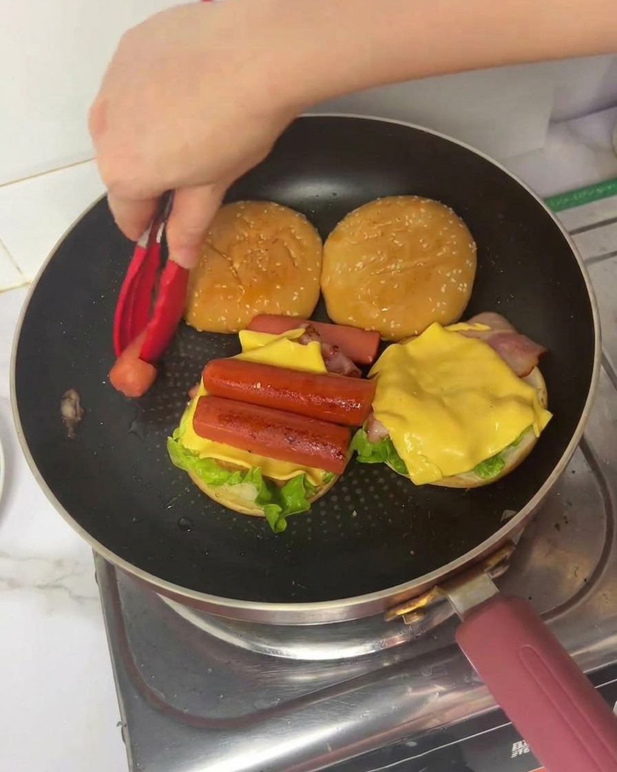 Una mano mette una porzione di frittata ripiegata su un burger stratificato con lattuga, bacon, formaggio e wurstel.