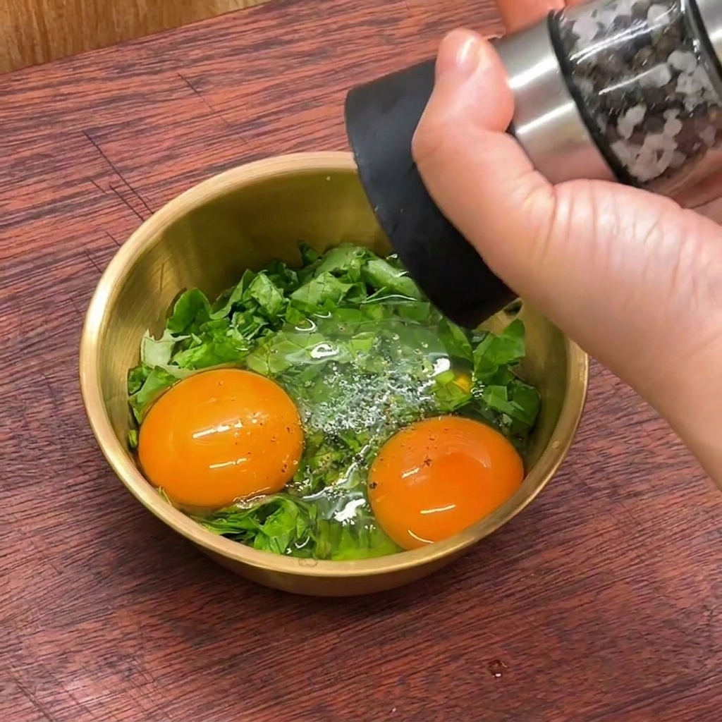 Una mano usa un macinapepe per condire due tuorli duovo e lattuga tritata in una ciotola dorata.