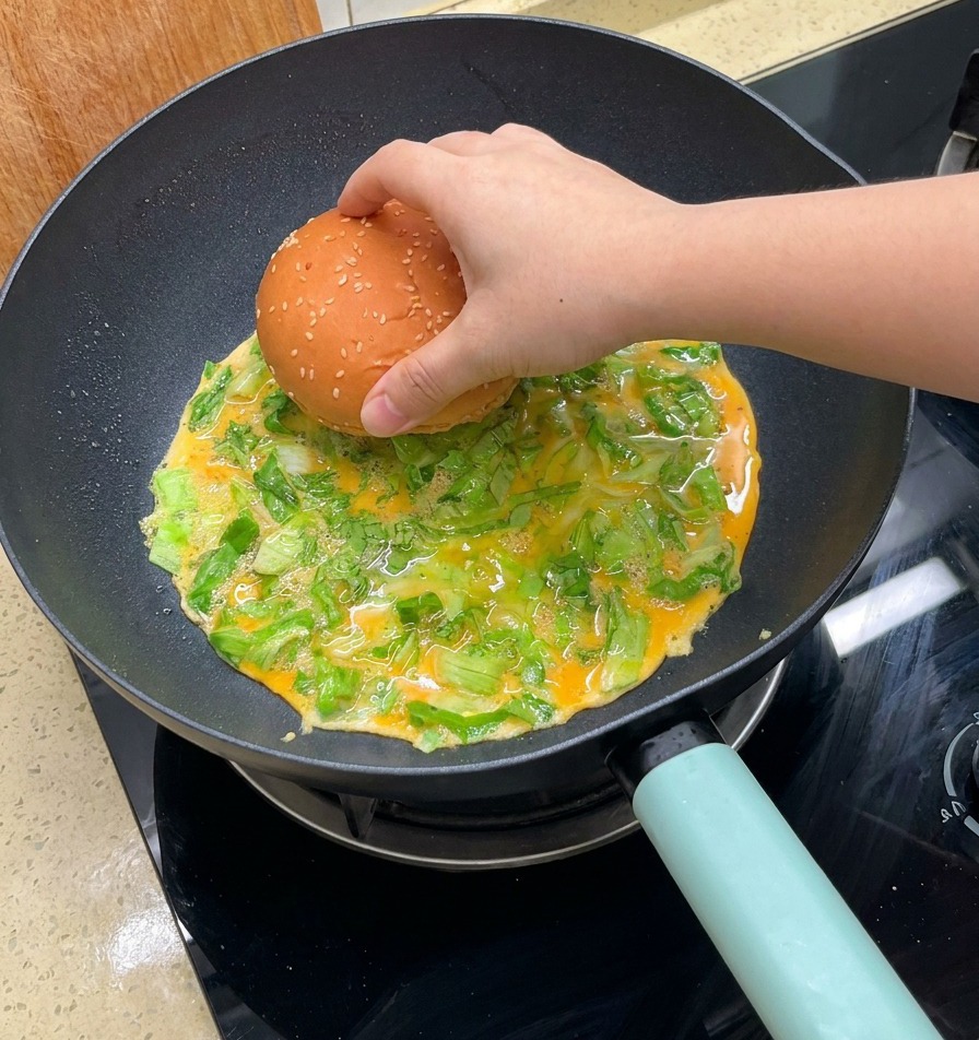 Una mano posiziona un panino al sesamo sulla frittata in cottura con lattuga in padella.