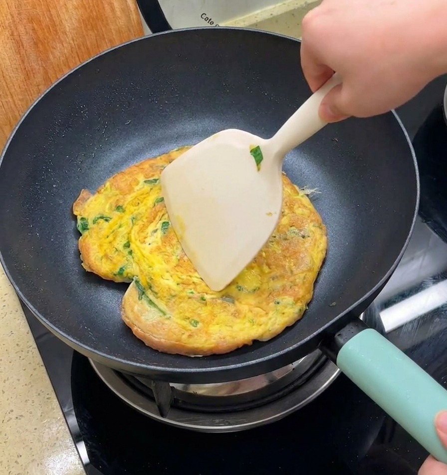 Una mano usa una grande spatola per girare una frittata dorata contenente verdure e pane in una padella nera.
