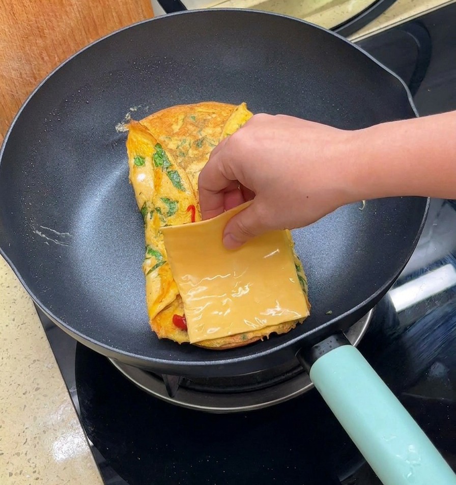 Una mano posiziona una fetta quadrata di formaggio giallo su una frittata ripiegata in padella.