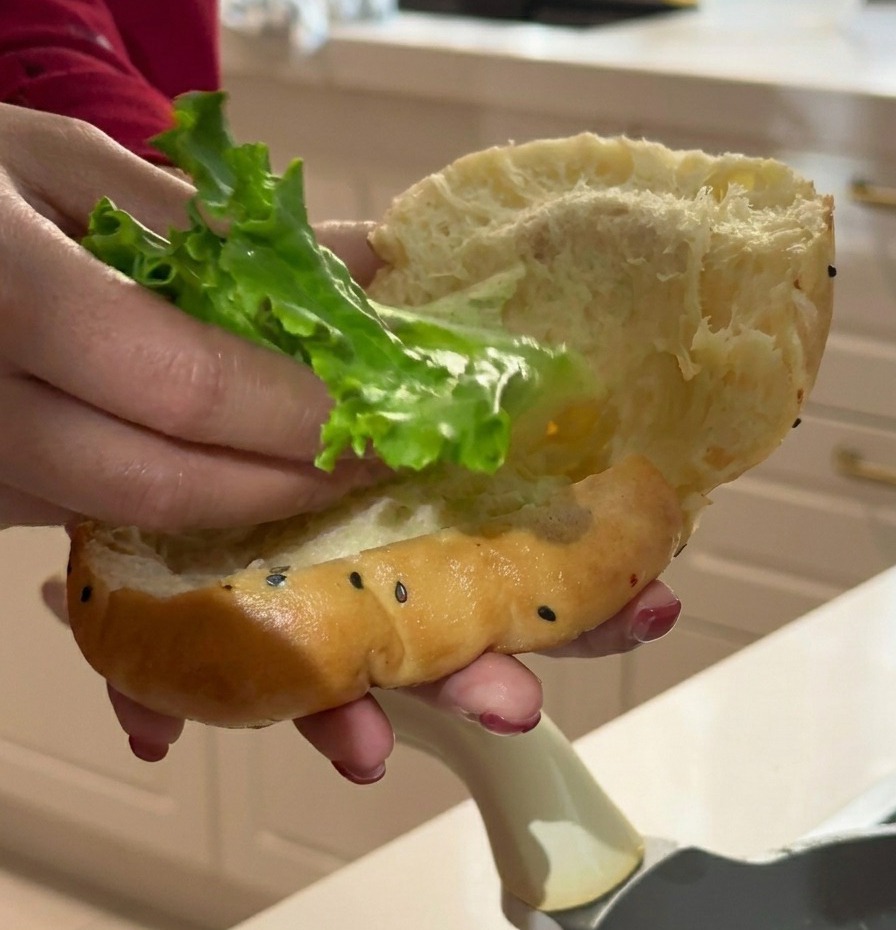 Mano che inserisce una foglia di lattuga fresca in un panino soffice ai semi di sesamo nero.