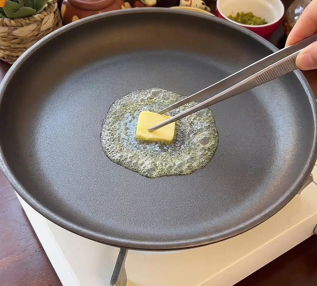 Due metà rotonde di panino per hamburger disposte a faccia in giù in una padella scura con burro fuso sfrigolante.