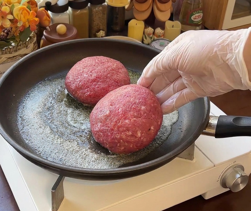 Due spessi e rotondi hamburger di manzo crudi inseriti in una padella con burro fuso, con una mano guantata visibile sulla destra.