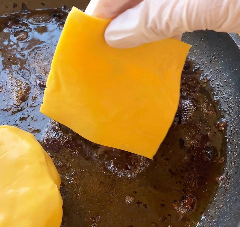Una mano guantata che posiziona una fetta di formaggio cheddar giallo intenso su un hamburger cotto che sfrigola in padella.