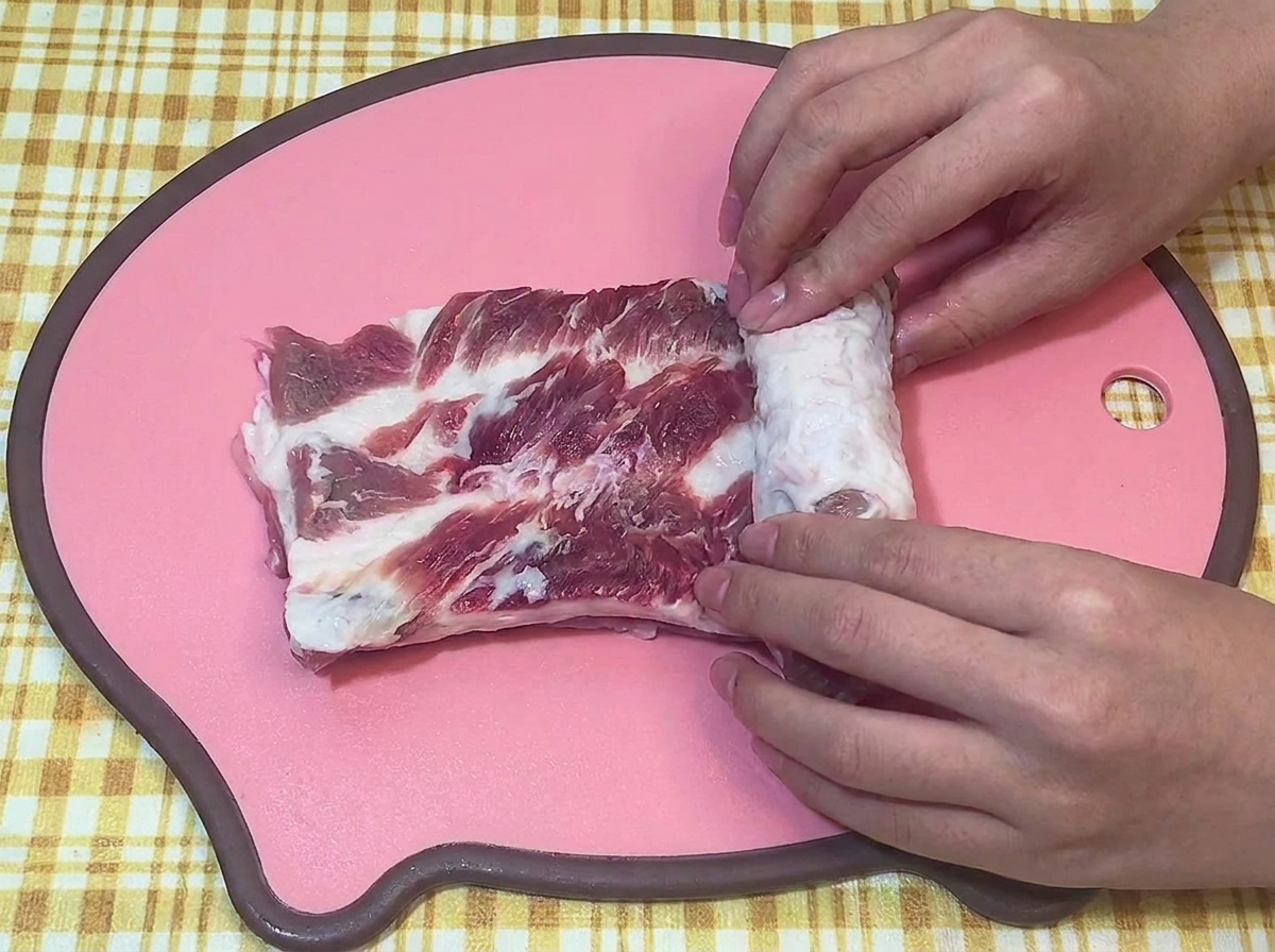 Mani che arrotolano strettamente un pezzo di pancetta di maiale cruda su un tagliere rosa.