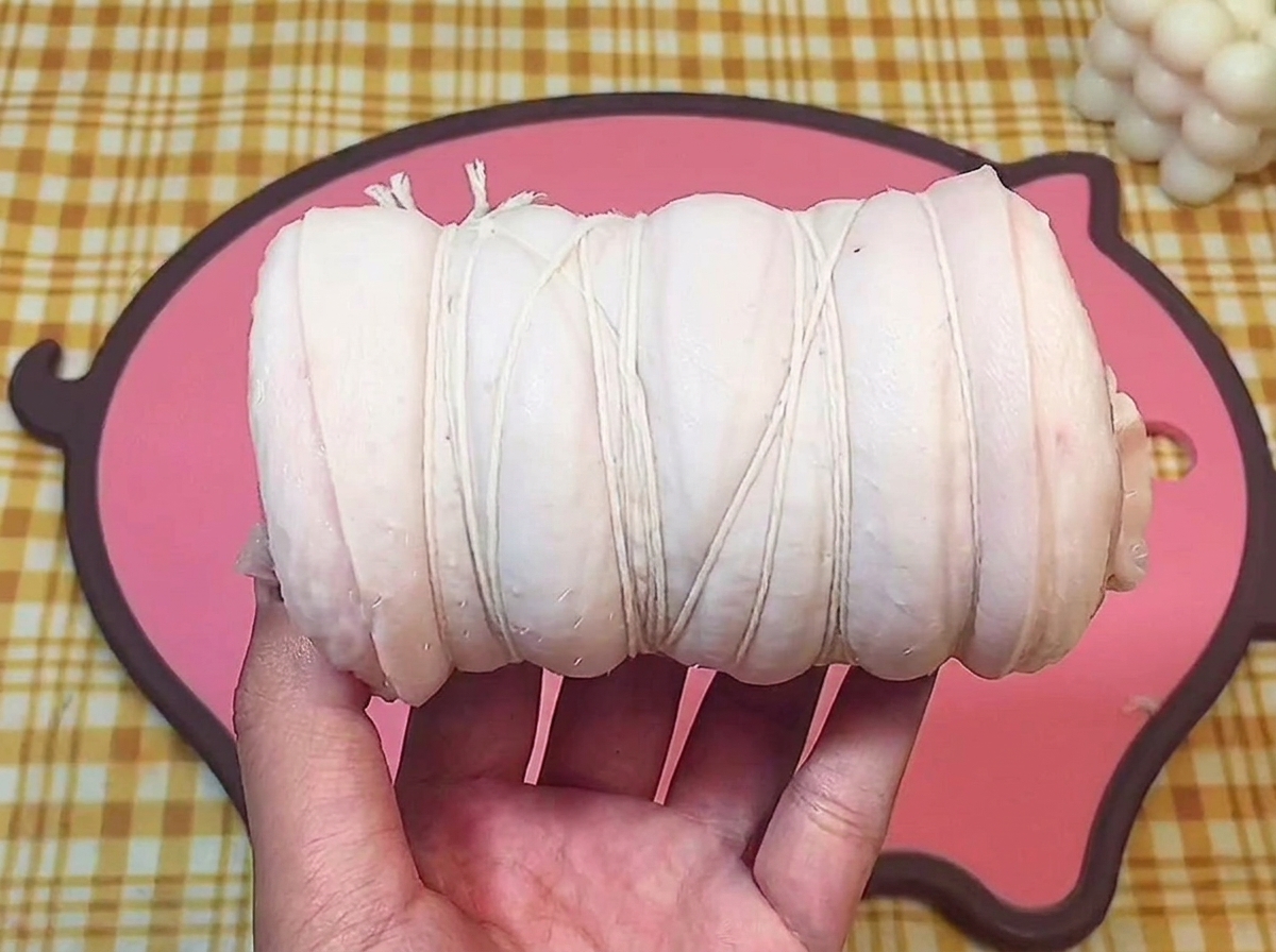 Una mano che tiene il rotolo di pancetta legato saldamente con spago da cucina bianco.