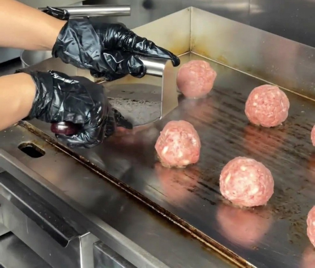 Primo piano di una pressa per hamburger in acciaio inossidabile che appiattisce una polpetta di manzo su una piastra commerciale.
