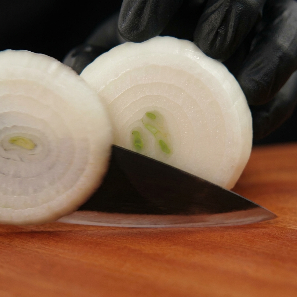 Mani con guanti neri che usano un coltello per tagliare una cipolla bianca sbucciata ad anelli su un tagliere di legno.