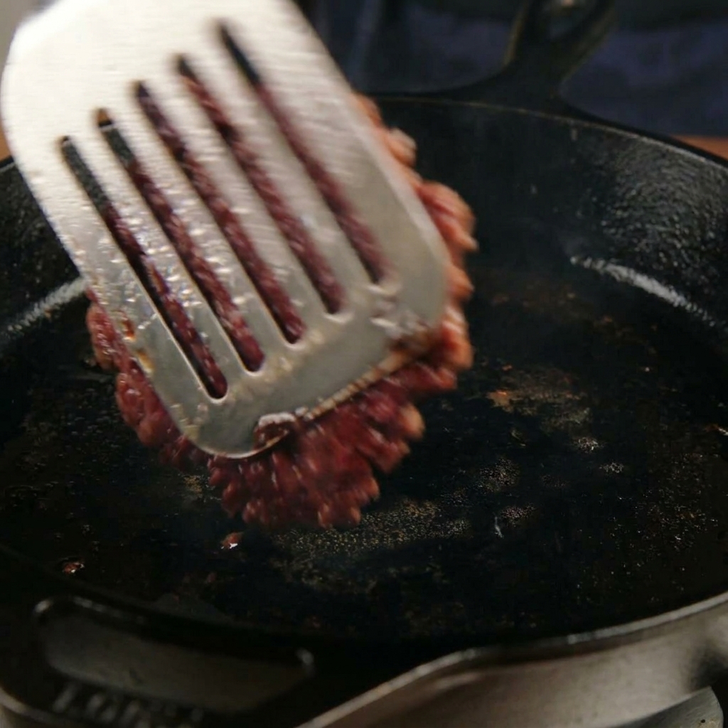 Girare un hamburger schiacciato e caramellato in padella con una spatola di metallo.