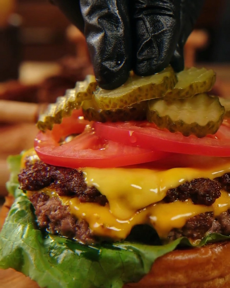 Dita con guanti neri che posizionano con cura fette di cetriolini ondulati su fette di pomodoro rosso vivo in cima a un cheeseburger impilato.