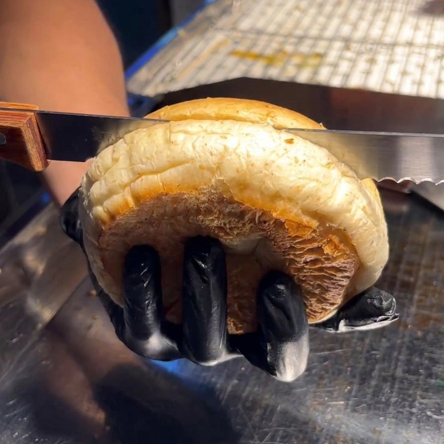 Una persona usa un coltello seghettato per tagliare a metà orizzontalmente un soffice panino da burger su una superficie dacciaio.