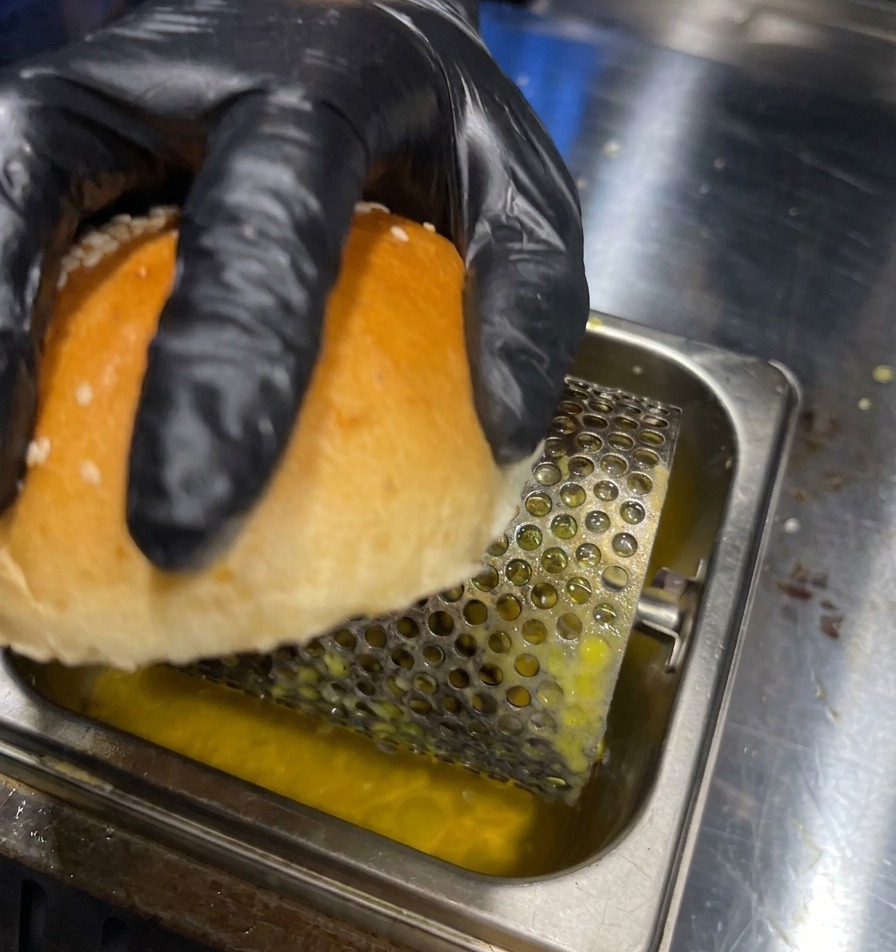 Una mano guantata fa rotolare il lato interno di un panino su un rullo per il burro in una vaschetta di metallo.