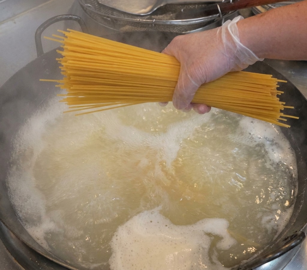 Una mano che indossa un guanto trasparente tiene un mazzetto di spaghetti secchi sopra una pentola di acqua bollente fumante.