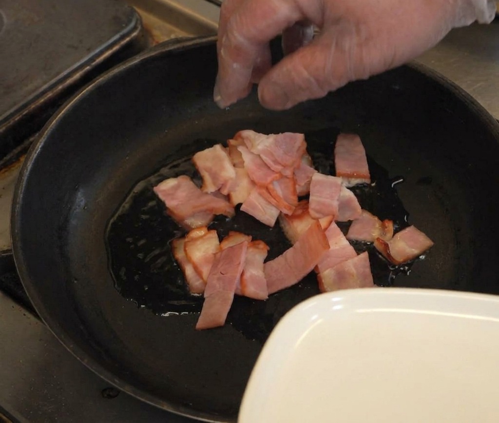 Una mano che mette fette di pancetta cruda in una padella scura con un velo dolio luccicante.