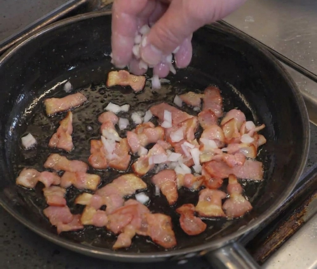 Una mano che sparge cipolla bianca tritata su strisce di pancetta parzialmente cotte in padella.