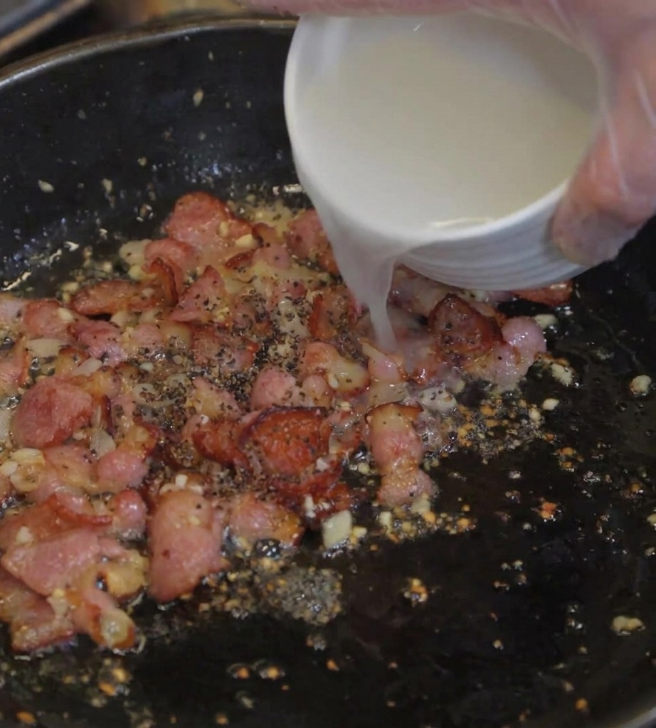 Acqua della pasta torbida che viene versata da una ciotola bianca in una padella con pancetta croccante e aromi.