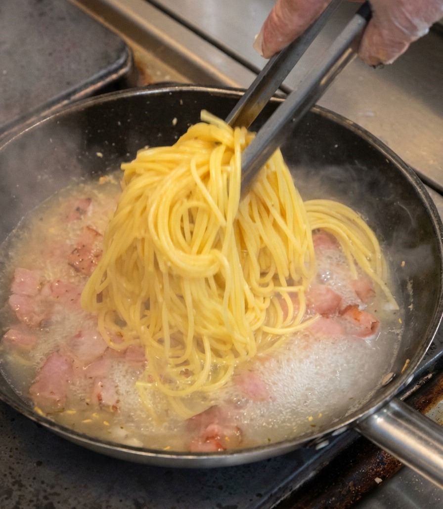 Un mazzetto di spaghetti gialli prelevato con le pinze e messo in una padella con pezzetti di pancetta e liquido bollente.