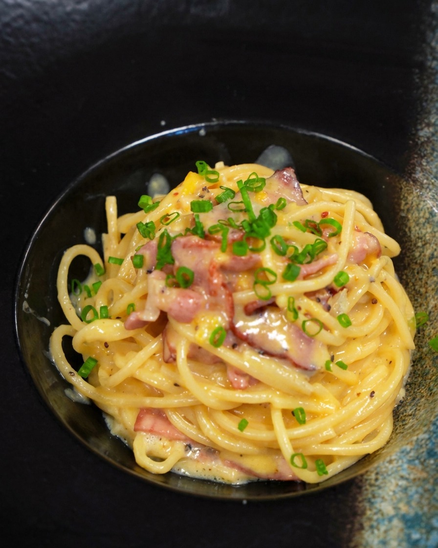 Una porzione di carbonara cremosa in una ciotola nera guarnita con una manciata di erba cipollina tritata.