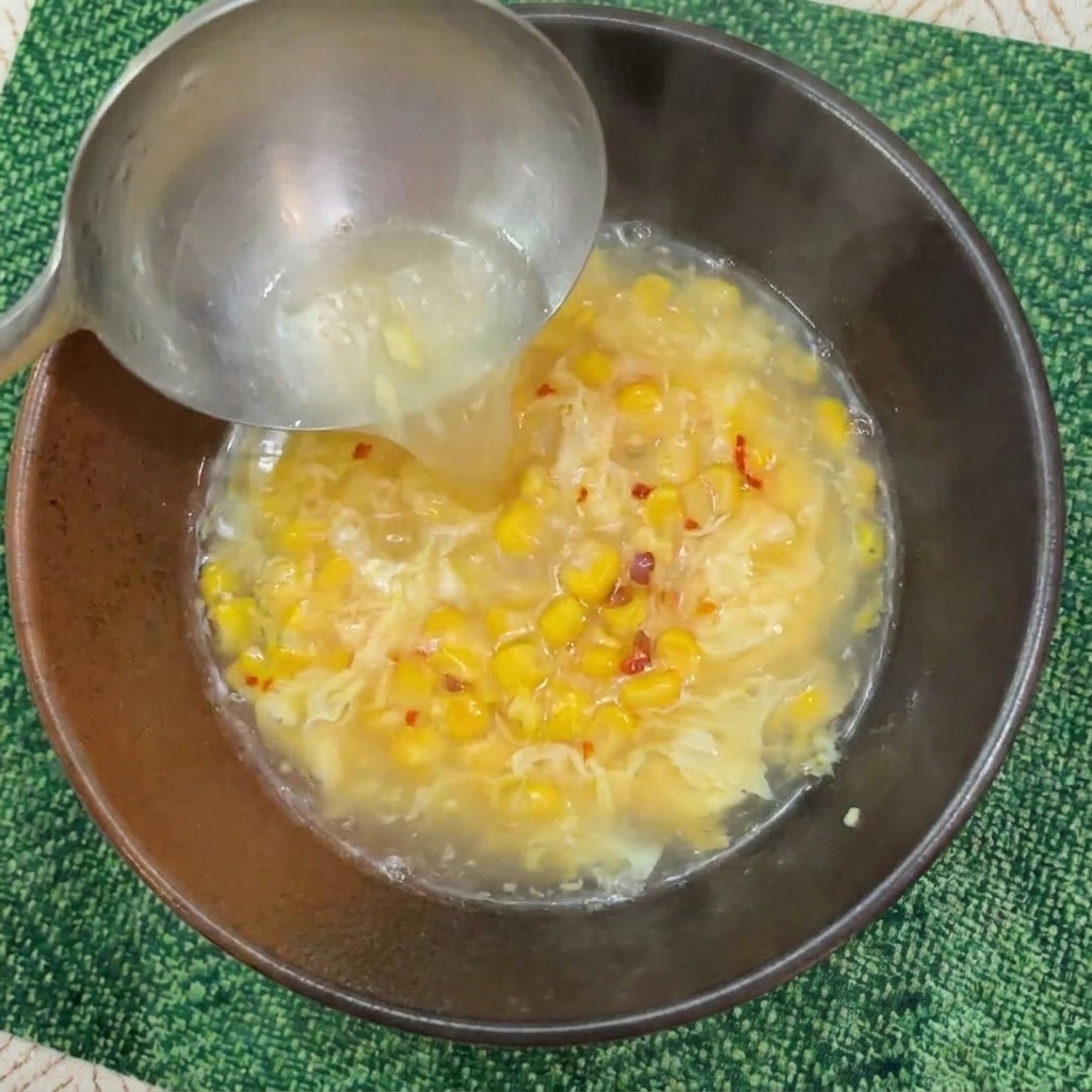 Un mestolo di metallo versa acqua bollente fumante in una ciotola con mais e il mix di uovo.